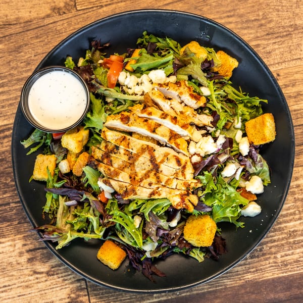 Grilled chicken salad with mixed greens, croutons, feta cheese, tomatoes, and a side of ranch dressing.