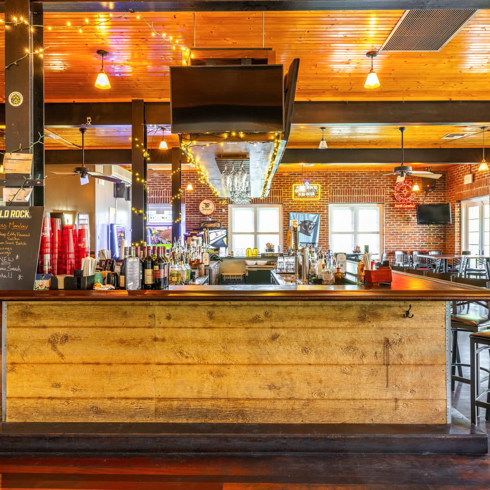 A rustic wooden bar with bottles, glasses, and a menu board, in a restaurant with brick walls.