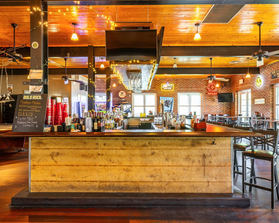 A rustic wooden bar with bottles, glasses, and a menu board, in a restaurant with brick walls.