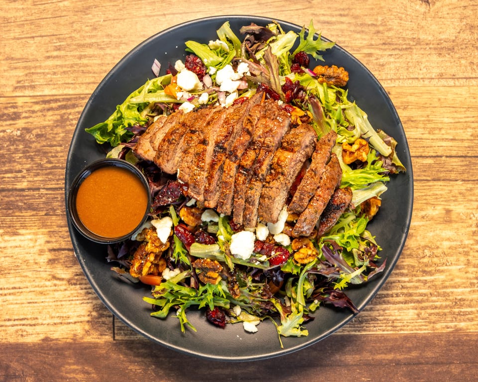 Grilled steak salad with mixed greens, feta, walnuts, cranberries, and a side of dressing.