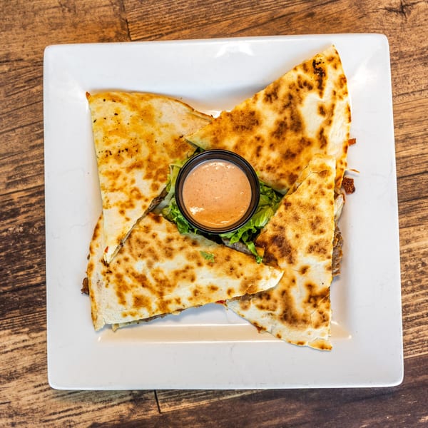 Four quesadilla wedges with a side of dipping sauce on a bed of lettuce, on a white plate.