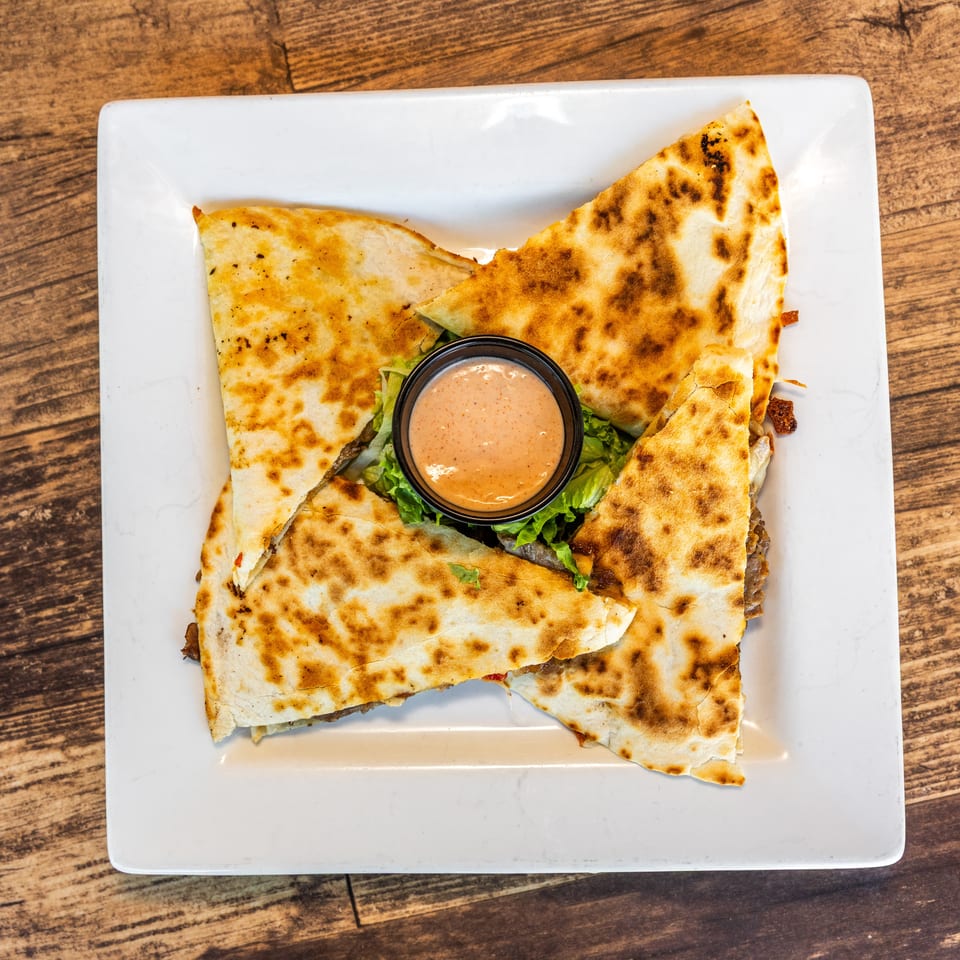 Four quesadilla wedges with a side of dipping sauce on a bed of lettuce, on a white plate.