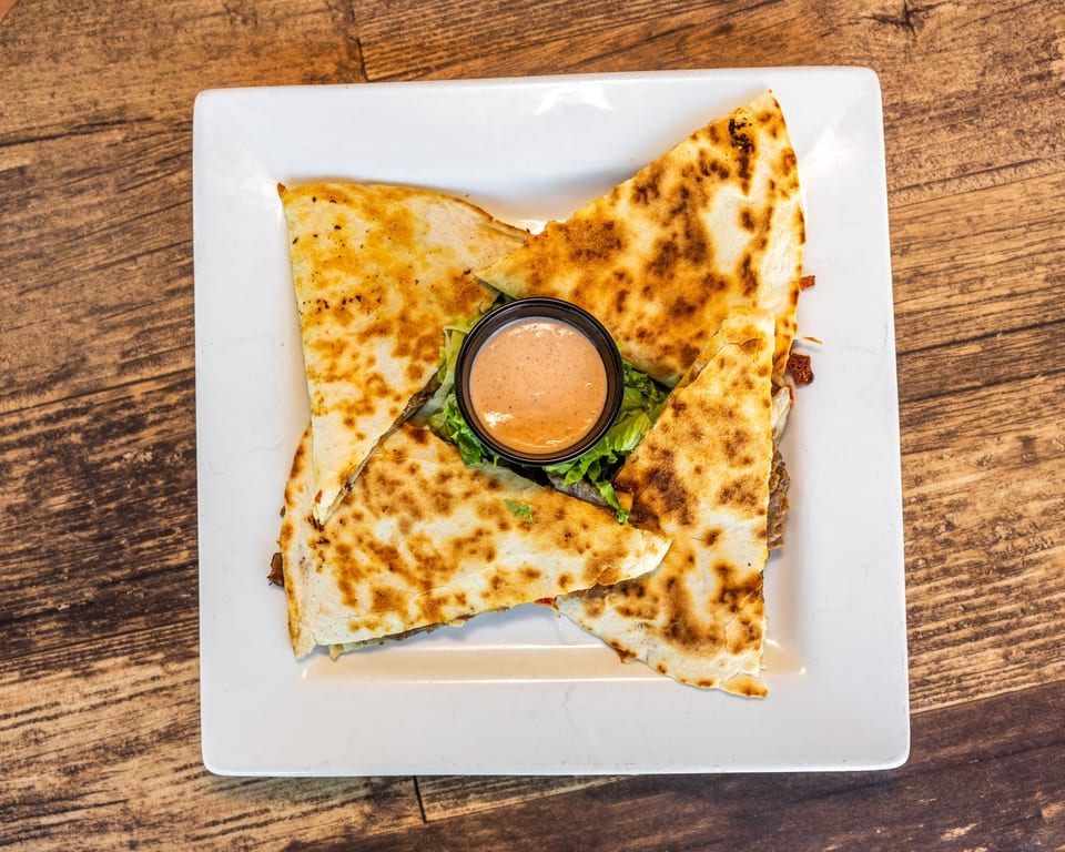 Four quesadilla wedges with a side of dipping sauce on a bed of lettuce, on a white plate.