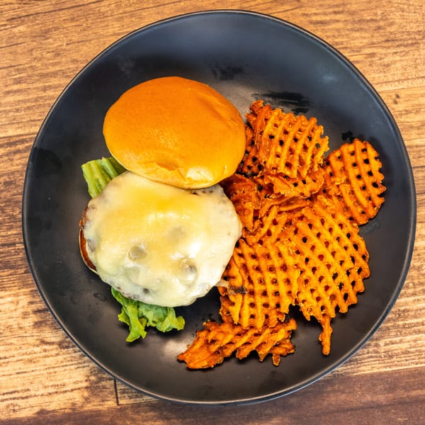 Cheeseburger with melted cheese and lettuce on a bun, served with waffle fries on a black plate.