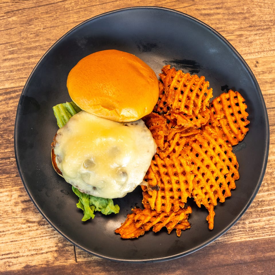 Cheeseburger with melted cheese and lettuce on a bun, served with waffle fries on a black plate.