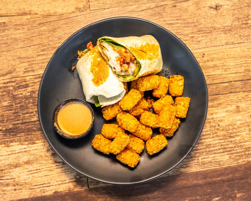A chicken wrap cut in half, tater tots, and a side of sauce on a black plate.