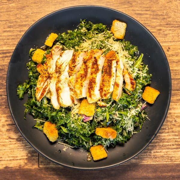 Grilled chicken Caesar salad with kale, croutons, and shredded cheese on a black plate.