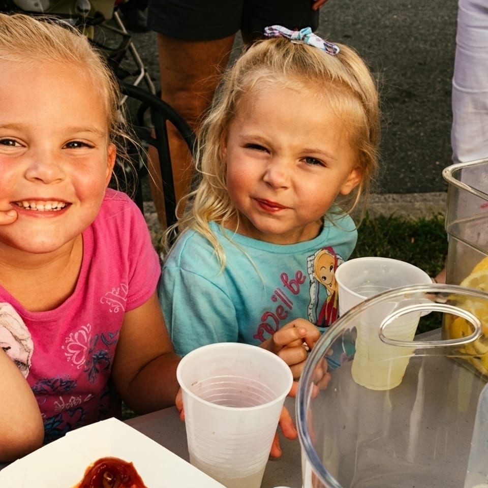 Two young blonde girls smiling at a table with lemonade and food outdoors.
