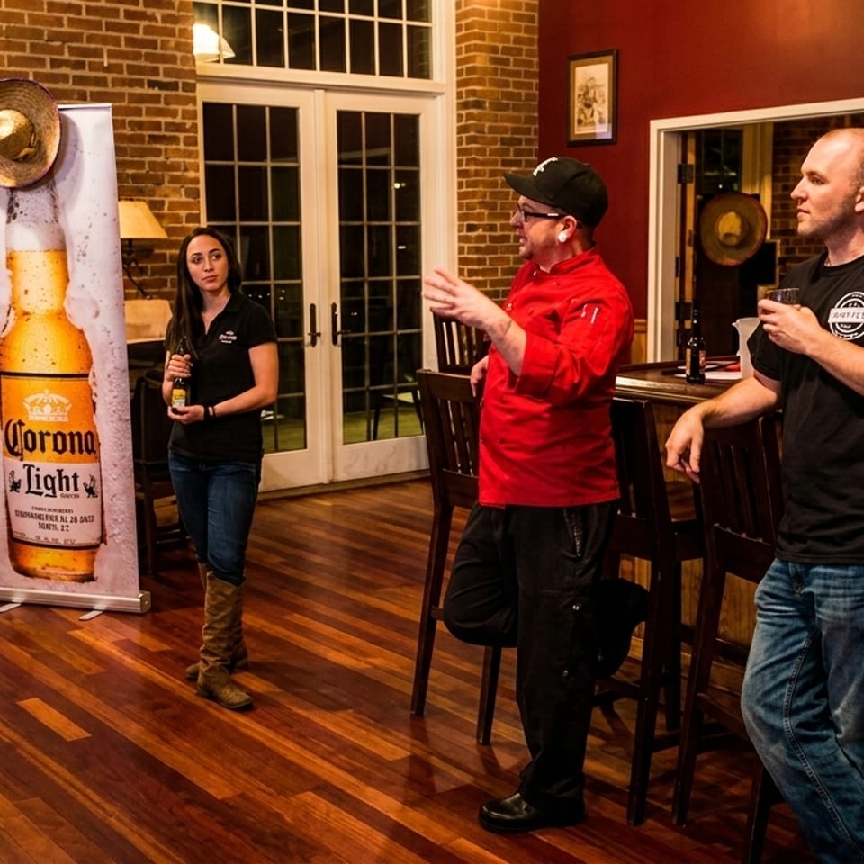 Three people, two men and one woman, stand and converse in a room with a Corona Light banner.