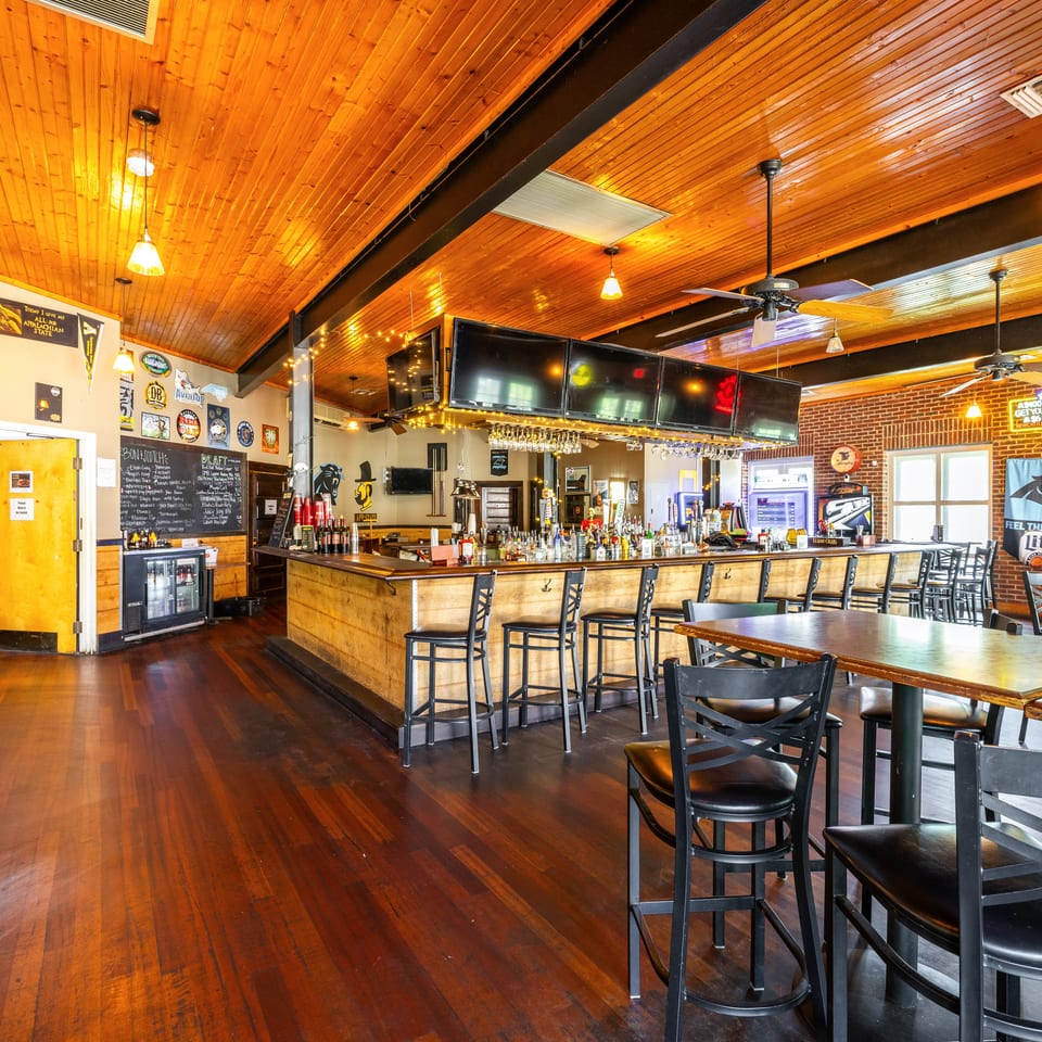 A rustic bar interior with a wooden bar, high-top tables, and a wooden plank ceiling.