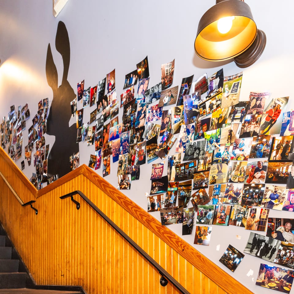 A staircase with wooden railings leads up to a wall covered in numerous framed photos and a large rabbit silhouette.