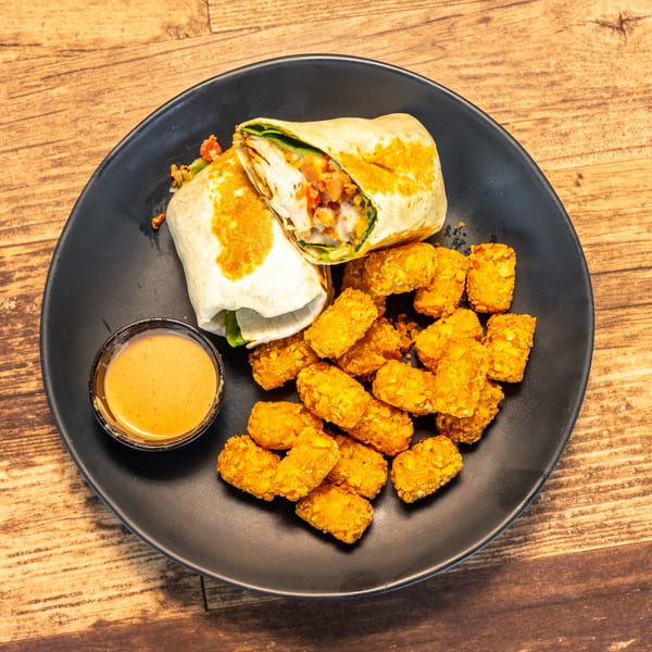 A chicken wrap cut in half, tater tots, and a side of dipping sauce on a black plate.